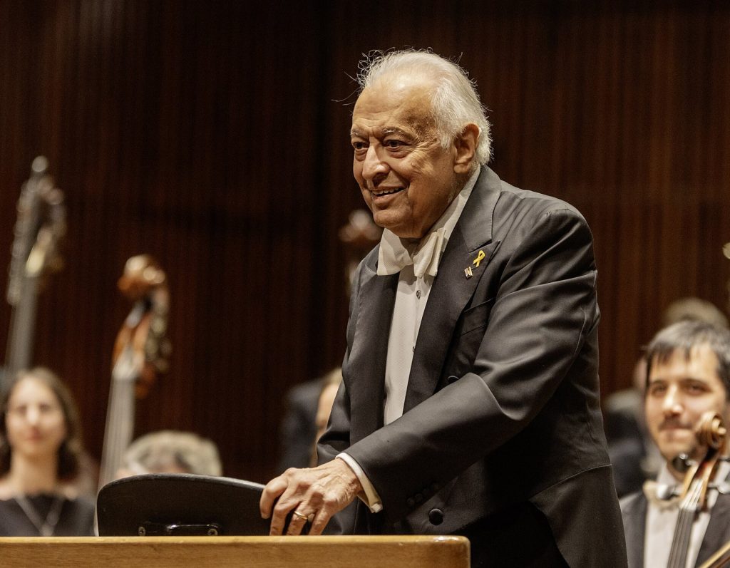 Zubin Mehta lächelnd am Dirigentenpult vor Orchestermusikern